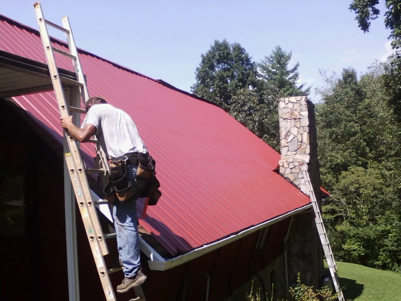 Roofer on a ladder beside a red metal roof and stone chimney — Soffit & Fascia Repair by Roofing Brothers in Natick